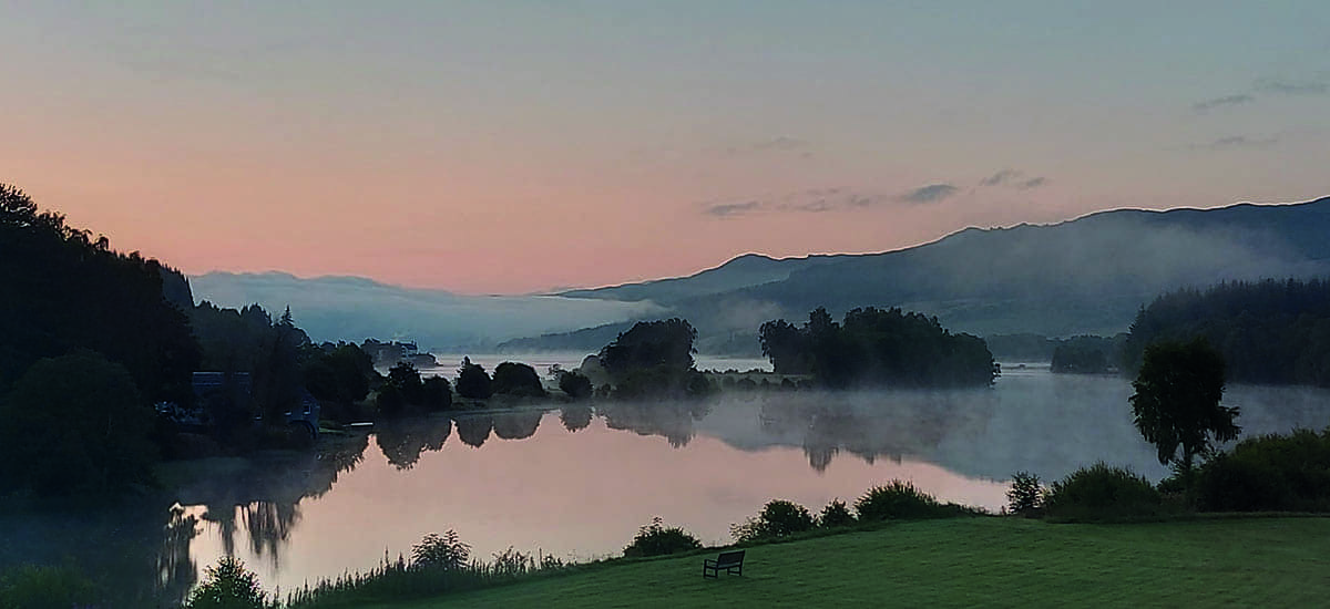 Sunrise over Loch Awe, with the sky bathed in warm hues of orange and pink reflecting on the tranquil waters and surrounding hills.