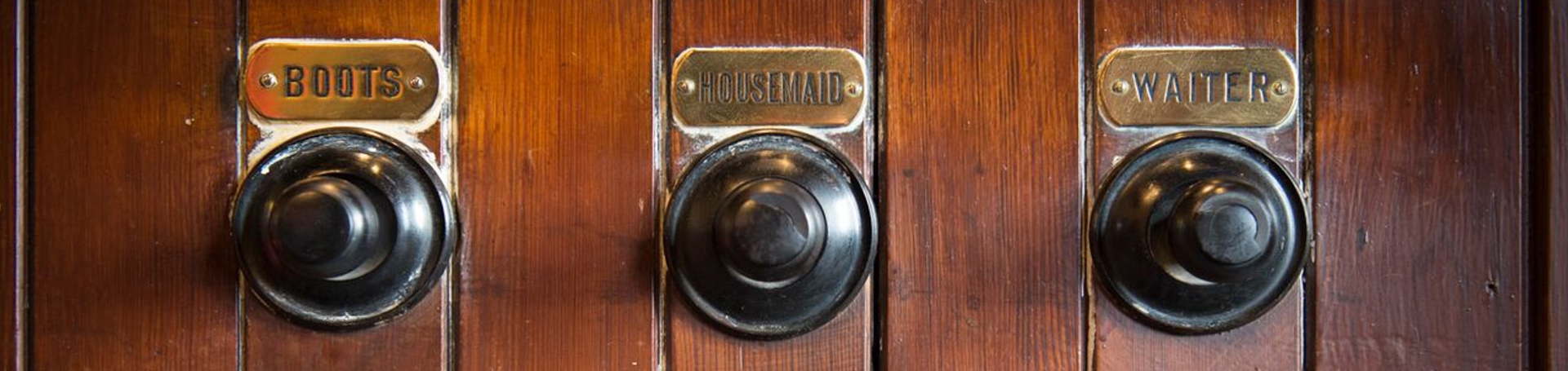 A image of a wooden panel with hangers and above each hanger a brass name plate with boots, housemaid, waiter.